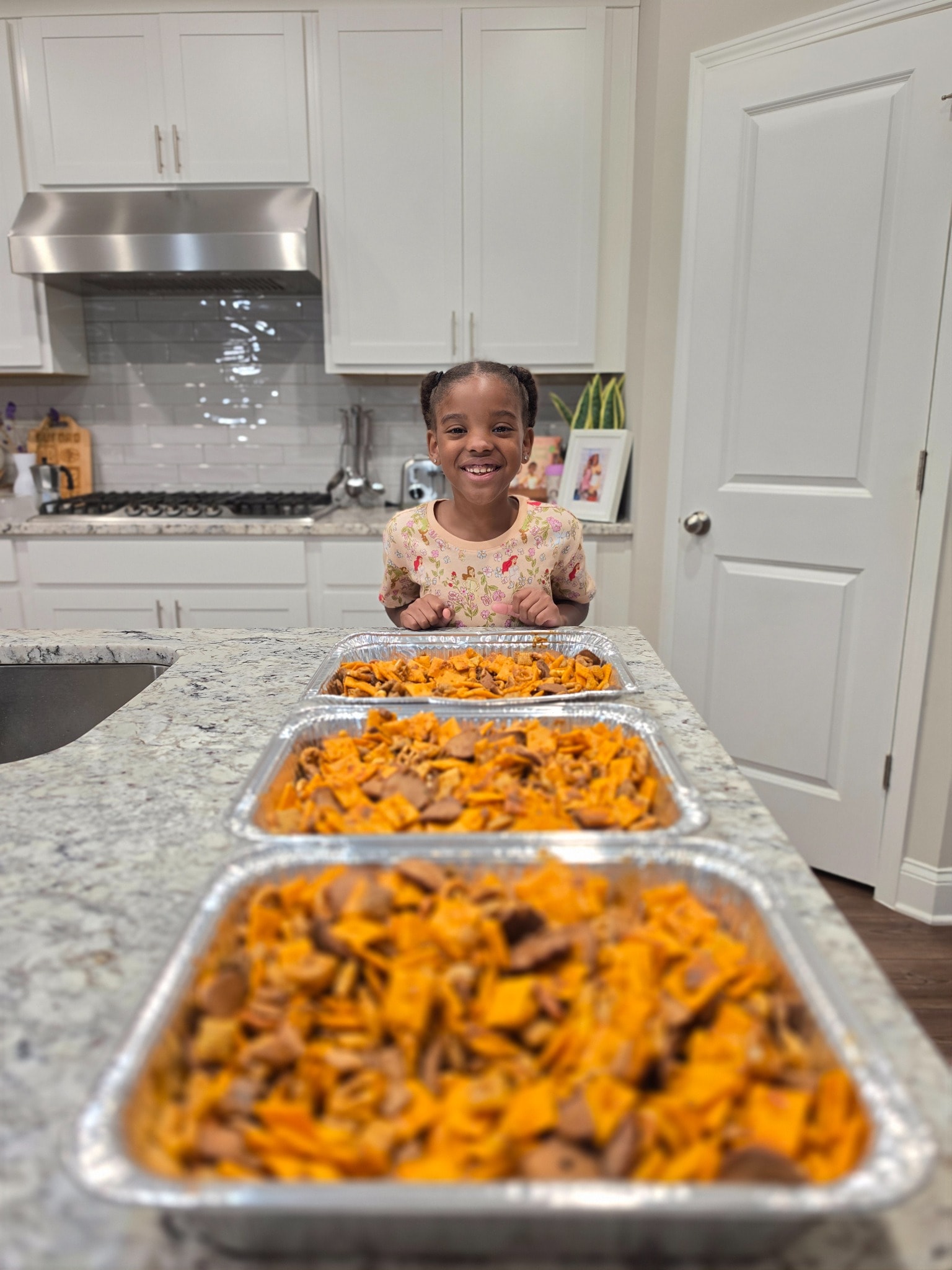 Luna smiling behind three trays of freshly made Luna & Dad Smokehouse snack mix in the family kitchen
