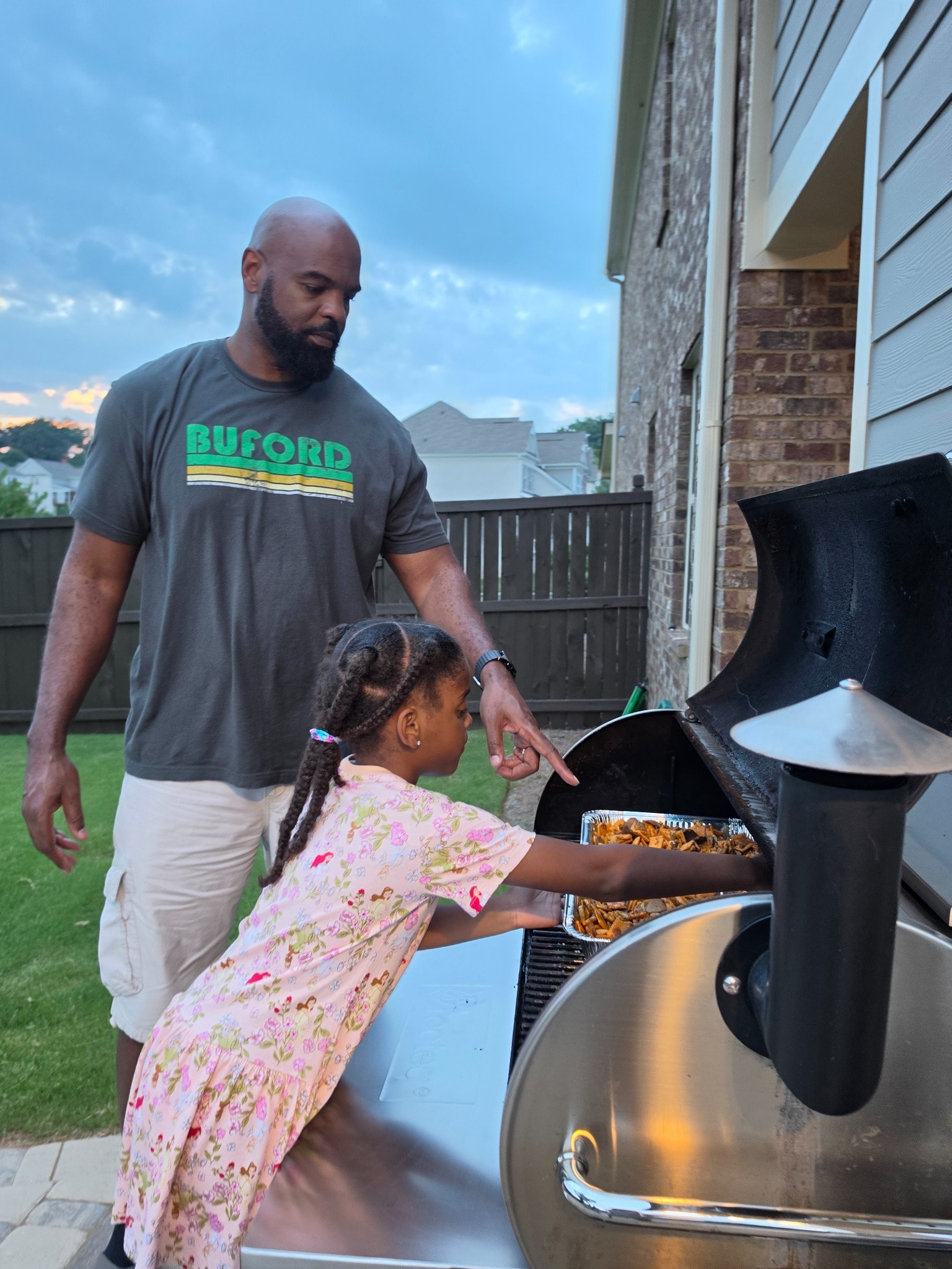 Dad pointing and guiding Luna as she slides a tray of snack mix into the smoker in the backyard in Buford, Georgia
